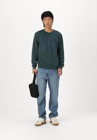 Pulover verde cu guler rotund, având logo-ul "GAP" în albastru închis, asortat cu blugi deschiși la culoare și adidași crem. Ține în mână o geantă neagră.