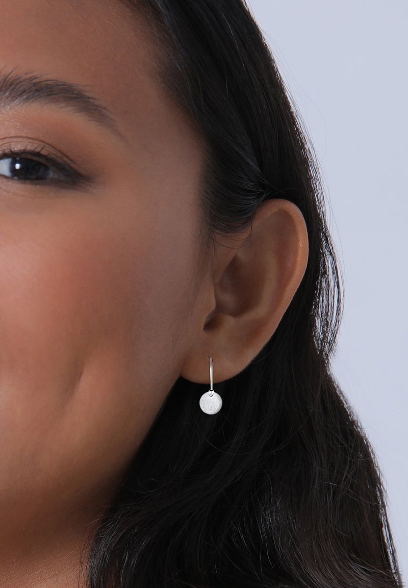 Pendientes de aro de plata con un charm circular texturizado, que exhiben un diseño minimalista. Los pendientes cuelgan justo por debajo del lóbulos de la oreja.