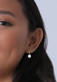 Srebrni obroči z teksturiranim okroglim obeskom. Kovina je videti polirana in odsevna, kar dopolnjuje temne, ravne lase.