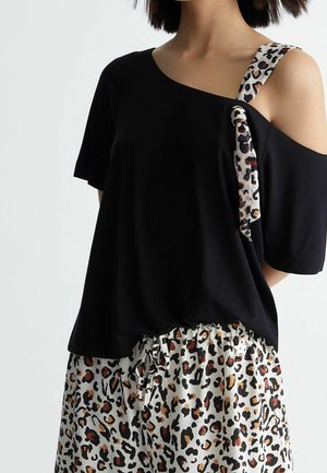 Kvinde iført sort off-shoulder top med leopardmønstret strop og matchende leopardmønstrede bukser med snøre i taljen.