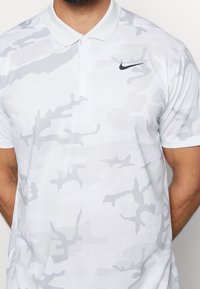 Muž v bílém a světle šedém maskáčovém polo tričku Nike s černým logem swoosh na levé části hrudi, stojící na bílém pozadí.