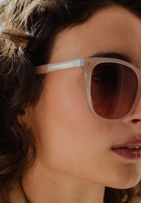 Sonnenbrille mit transparenten Kunststoffrahmen und dunkelbraunen Gläsern. Merkmale sind ein Logo am Bügel und ein modernes quadratisches Design.