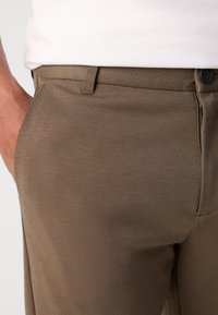 Pantalones de tela trenzada marrones con una textura suave, que presentan un bolsillo lateral y una bragueta con cremallera, complementados con un botón negro.