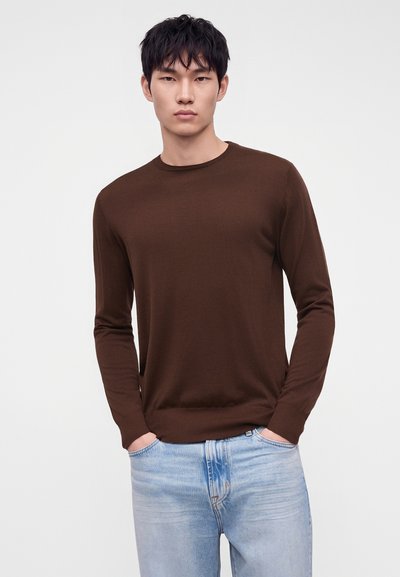 Jeune homme aux cheveux courts et noirs portant un pull marron uni à manches longues et un jean bleu clair, les mains dans les poches.