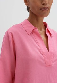 Rosa Hemd mit Kragen, kurzen Ärmeln und V-Ausschnitt. Hergestellt aus einem leichten Stoff mit einer glatten, strukturierten Oberfläche.