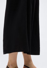 Kājas personas, kas valkā garu melnu svārku un melnas sandales ar siksniņām uz vienkārša gaiša fona.