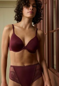 Vrouw met krullend haar in een bijpassende bordeauxrode beha en hooggesneden kanten panties, staande bij een houten raamkozijnen.