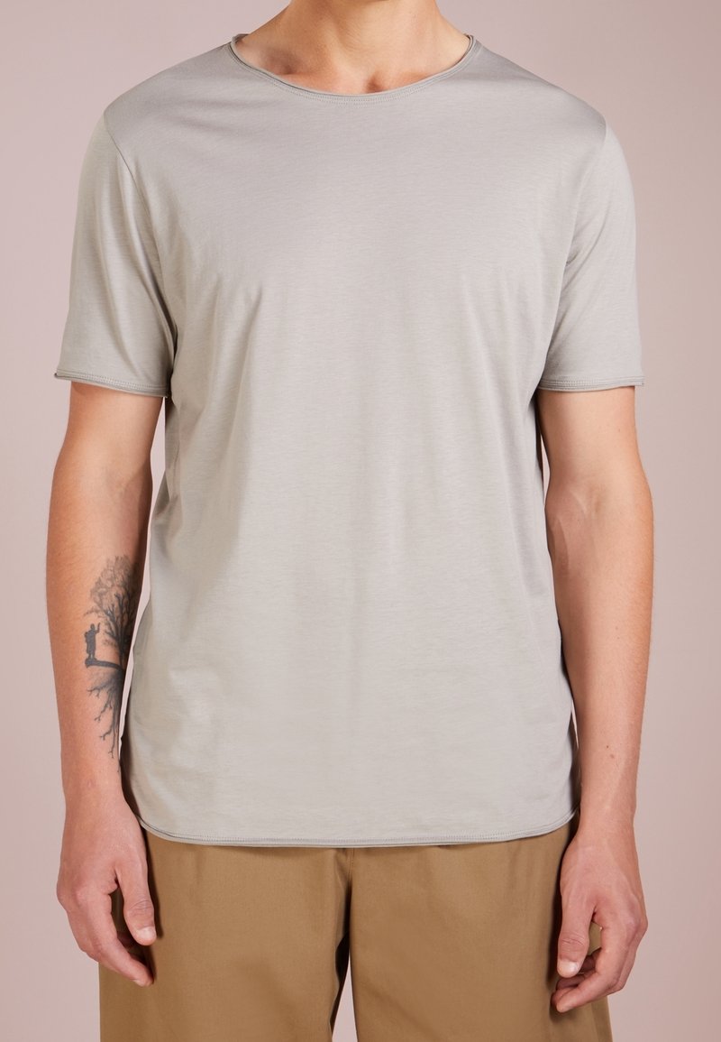 Lichtgrijs T-shirt met korte mouwen, ronde hals, zachte textuur, glad materiaal, gedragen met beige broek, met een kleine tatoeage op de arm.