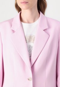 Blazer rosa claro con cuello con muesca, cierre de un botón y tela suave. Llevado sobre una camisa blanca con un patrón y pedrería.