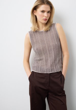 Mujer con un top de punto sin mangas con rayas verticales y pantalones marrón oscuro, de pie con las manos en los bolsillos frente a una pared gris claro.