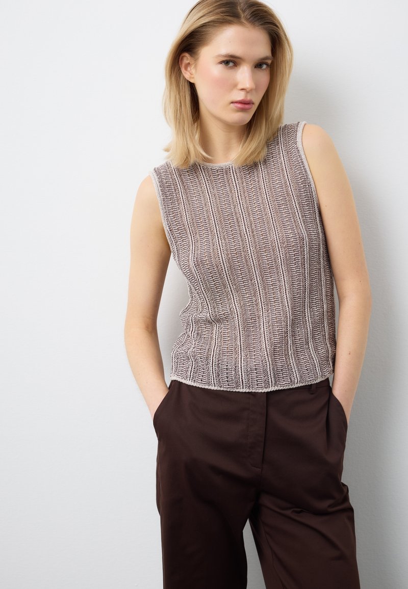 Frau trägt ein ärmelloses Strickoberteil mit vertikalen Streifen und dunkelbraune Hose, steht mit den Händen in den Taschen vor einer hellgrauen Wand.