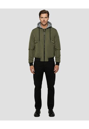 Zielona kurtka typu bomber z szarym kapturem, zapinana na zamek, z prążkowanymi mankietami, zestawiona z czarnymi spodniami i czarnymi butami. Gładka tekstura, swobodny styl.