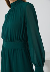 Robe teal en tissu léger et transparent avec un col haut, un corsage froncé et de longues manches légèrement bouffantes. Détail de taille élastique.