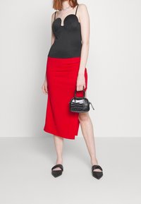 Schwarzes, tailliertes Oberteil mit geschwungenem Ausschnitt, roter Midirock mit seitlichem Schlitz, schwarze lackierte Mini-Handtasche und schwarze spitze Ballerinas.