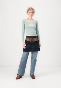 Cămașă scurtă cu mânecă lungă, de culoare verde deschis, cu textul "1998 SAN DIEGO". Asortată cu un kilt cu carouri albastre și o curea maro cu accente circulare. Blugi albastre și pantofi maro.