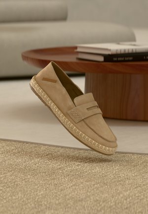 Beige mokasinsko i semsket skinn med vevd såle i espadrillestil, plassert i en stue i nøytrale toner nær et trebord med bøker.