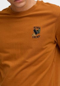 Brun bomulds t-shirt med et lille broderet mærke, der viser en illustration af en støvle, en banan og teksten "BORN LUCKY" i sort.