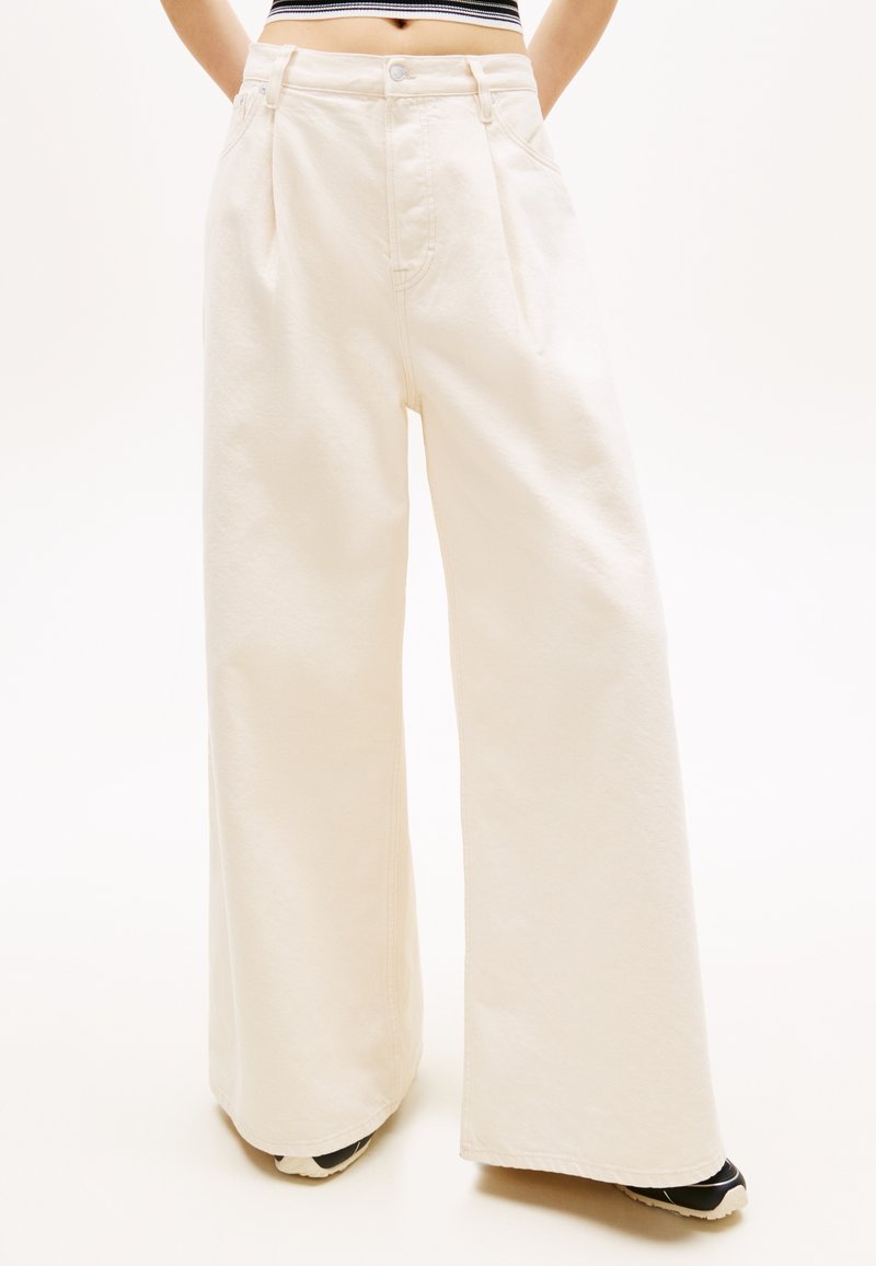 Ψηλός άνθρωπος που φοράει κρεμ παντελόνι υψηλής μέσης και με φαρδιά πόδια, καθώς και μπεζ και μαύρα αθλητικά παπούτσια, με τα χέρια στις τσέπες, το κάδρο κόβεται στη μέση.