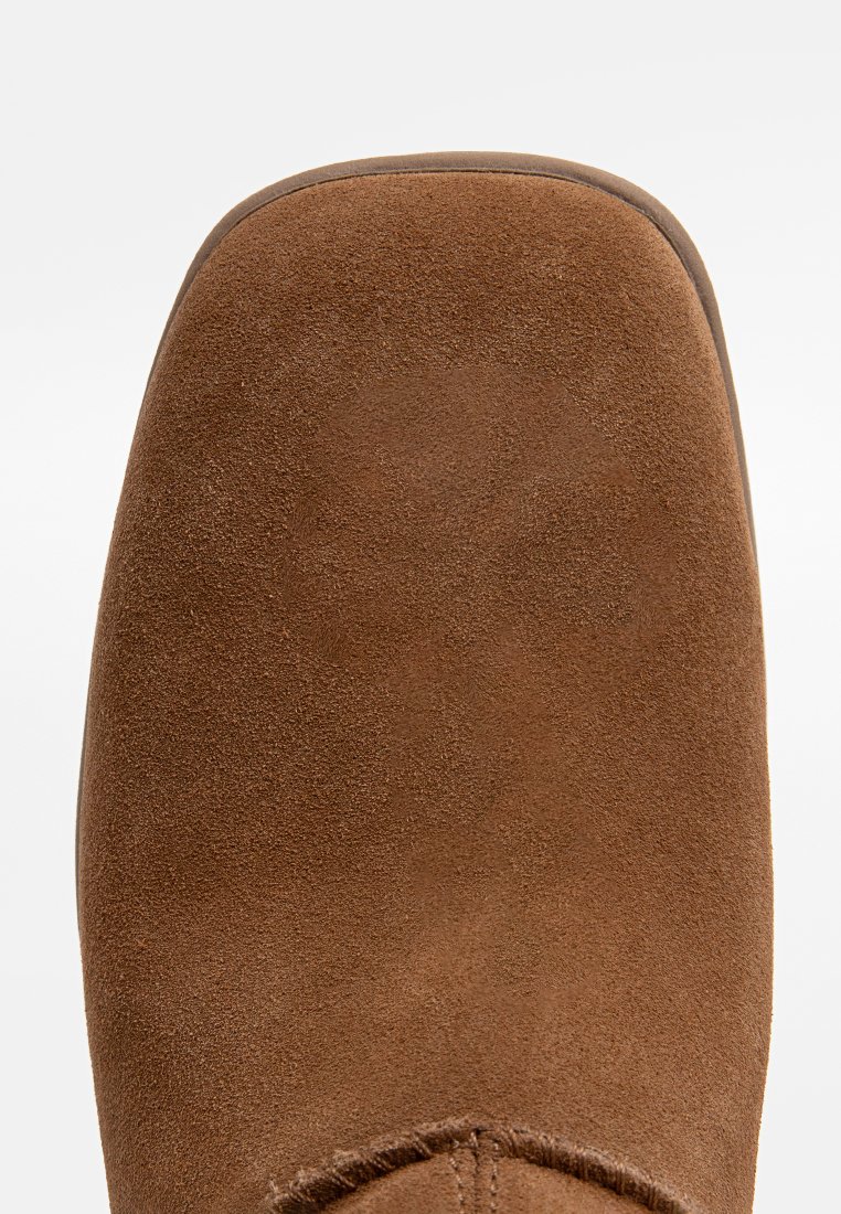 Bottine en daim marron avec un bout arrondi, des coutures visibles et une finition lisse et texturée. Design subtil avec une apparence douce.