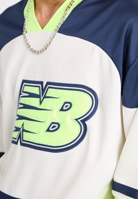 Valkoinen ja tummansininen verkkomateriaali, jossa on suuri neonvihreä "NB" -logo. Sisältää syvän V-pääntien ja lyhyet hihat, joissa on tekstuuripinta ja ketjukaulaketju.