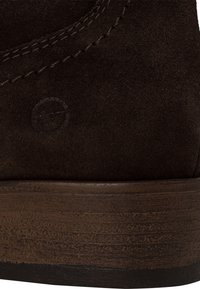 Brązowy zamszowy botek za kostkę z teksturowaną, ciemno drewnianą podeszwą i widocznymi detalami stworzonymi ze szwów. Okrągłe logo wytłoczone na bocznej części.
