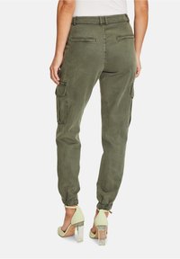 Zielone spodnie cargo z elastycznymi mankietami, posiadające boczne kieszenie, luźny krój oraz kontrastowe przeszycia. Widoczne przezroczyste buty na obcasie.
