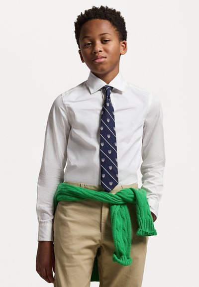 Camisa blanca, corbata azul marino con patrón de escudo, pantalones caqui, suéter verde de punto trenzado anudado a la cintura. Look casual y en capas.