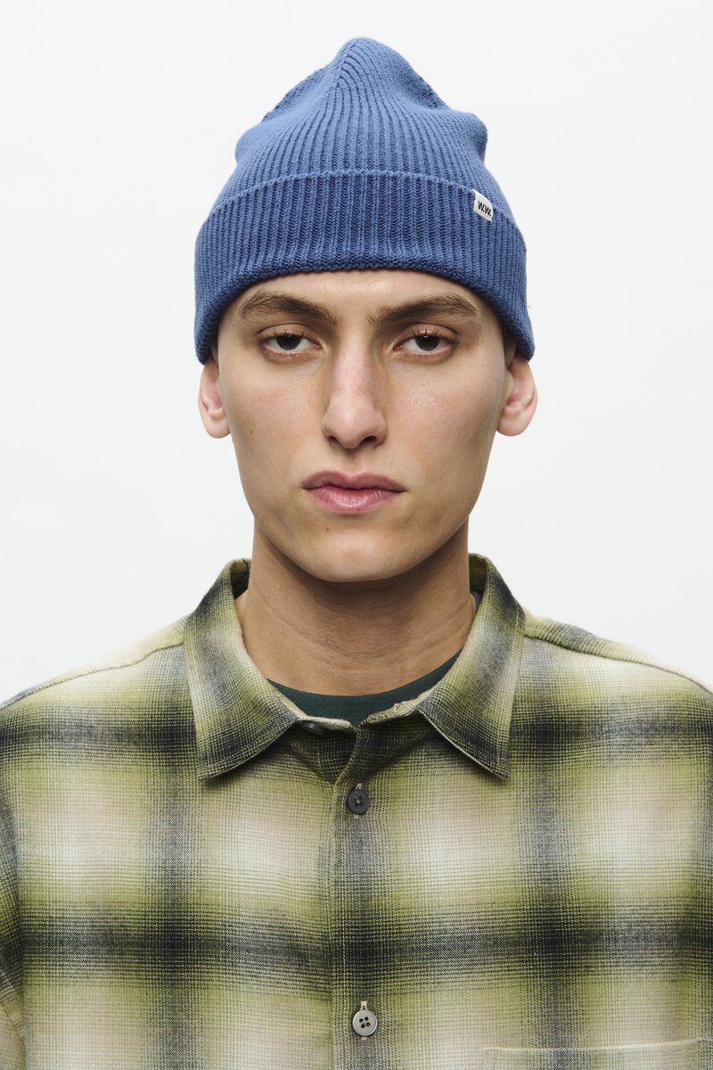 Sinine ribitud beanie terava tipuga ja väikese sildiga. Allpool on roheline ja must ruuduline flanellist särk nööbiklappidega.