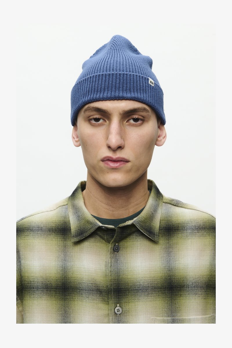 Blue ribbed beanie with a pointed top and a small label. Below, a green and black plaid patterned flannel shirt with button closure.