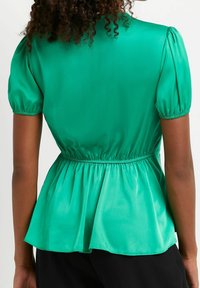 Grön satinpeplumtop med korta puffärmar, elastisk midja och mjuk textur. Tyget har en lätt lyster.
