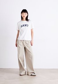 Balta marškinėliai su „GANT“ mėlynomis raidėmis, derinami su šviesiais plačiaaturių kelnių. Modelis stovi ant šviesaus grindų paviršiaus.