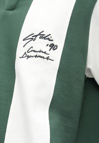 Groen-wit gestreepte sweatshirt met een polo-nek, voorzien van geborduurde tekst in marineblauw. Zacht katoenmateriaal met een gladde textuur.