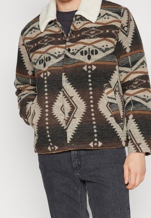 Mand iført en brun, beige og sort mønstret jakke med en hvid plyskrave og sorte jeans, hånd i jakkelommen.