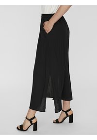 Schwarzer Midirock mit elastischem Bund, Seitentasche und mittellangem Schlitz. Kombiniert mit schwarzen Sandalen mit Blockabsatz. Glatte Textur.