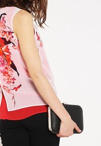 Sleeveless top with a sheer pink floral design, layered over a red shirt. The person holds a black textured clutch purse.