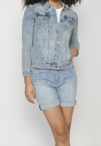 Vaaleansininen farkkutakki, jossa on kukkakirjontaa, napit ja kaksi rintataskua, yhdistettynä vastaaviin kirjottuihin farkkushortseihin.