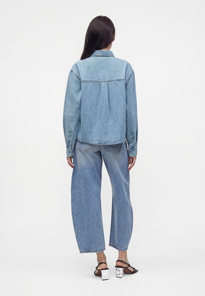 Femme aux longs cheveux foncés portant une chemise en denim clair, un jean à jambes larges et des sandales à talons noires, debout de dos sur un fond uni.