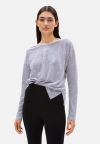 Frau mit langen dunklen Haaren, die ein langärmliges navy- und weiß gestreiftes Oberteil trägt, das in eine hochtaillierte schwarze Hose gesteckt ist, steht vor einem schlichten Hintergrund.
