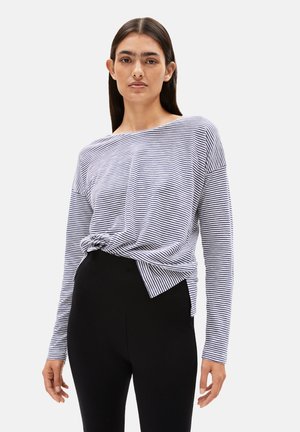 Vrouw met lang donker haar, gekleed in een navy en witte gestreepte top met lange mouwen, ingestopt in hoge zwarte broeken, staande tegen een effen achtergrond.