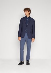 Navy blazer with two buttons, contrasted by a navy polka dot shirt. Paired with light blue patterned trousers and black shoes.