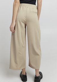 Beige laiud püksid elastse vöökohtaga, millel on üks tagatasku ja sile tekstuur, koos mustade platvormkingadega.