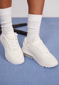 Vita atletiska sneakers med meshpaneler, snörning och en genomskinlig dämpningssula. I kombination med vita ankelsockar på en blå yta.