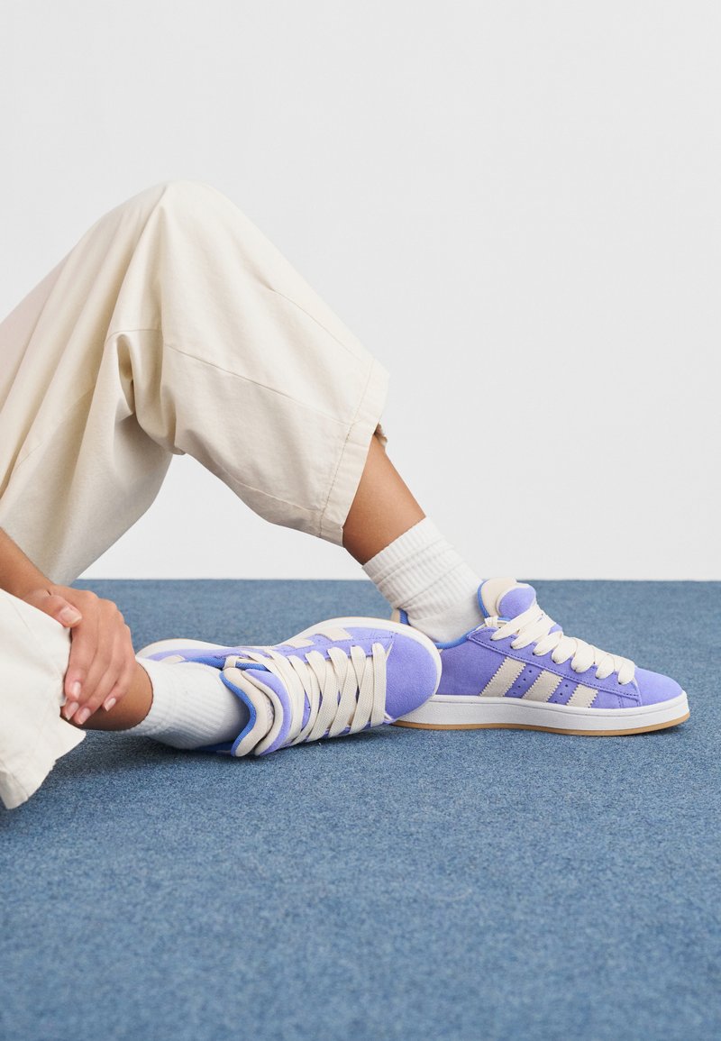 Purple suede sneakers with cream-colored laces and stripes. Features a rubber sole and blue accents. Worn with white crew socks and beige pants.