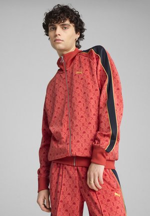 Rød zip-up træningsjakke med mønstret tekstur, ribbet krave og manchetter. Sorte og gule striber løber langs ærmerne.