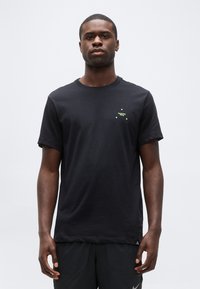 Νεαρός μαύρος άνδρας στέκεται, φορώντας μαύρο γραφικό μπλουζάκι με μικρό κίτρινο σχέδιο στο στήθος και μαύρο σορτς, απέναντι σε απλό φόντο.