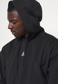 Zwarte hoodie gemaakt van zachte stof, met een klein wit Adidas-logo op de borst. Het ontwerp bevat een trekkoord voor verstelbaarheid.