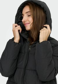 Melnā pūkādā jakā ir rāvējslēdzēja priekšpuses aizdare, elastīgas aproces un kapuce. Materiāls izskatās gluds, ar kviltētām daļām siltumizolācijai.