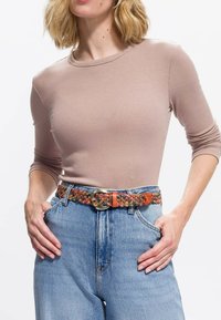 Kobieta w be�żowej, prążkowanej bluzce z długim rękawem, włożonej w wysokiej talii niebieskie dżinsy, z kolorowym, plecionym skórzanym paskiem i rękami w kieszeniach.