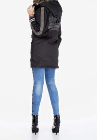Parka nera con cappuccio, caratterizzata da dettagli argentati e design ricamato sul retro. Abbinata a jeans skinny blu chiaro e tacchi alti neri.