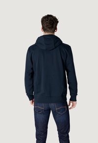 Felpa con cappuccio di colore blu navy con texture liscia, polsini e vita a costine, indossata con jeans blu scuro. Solo vista posteriore.